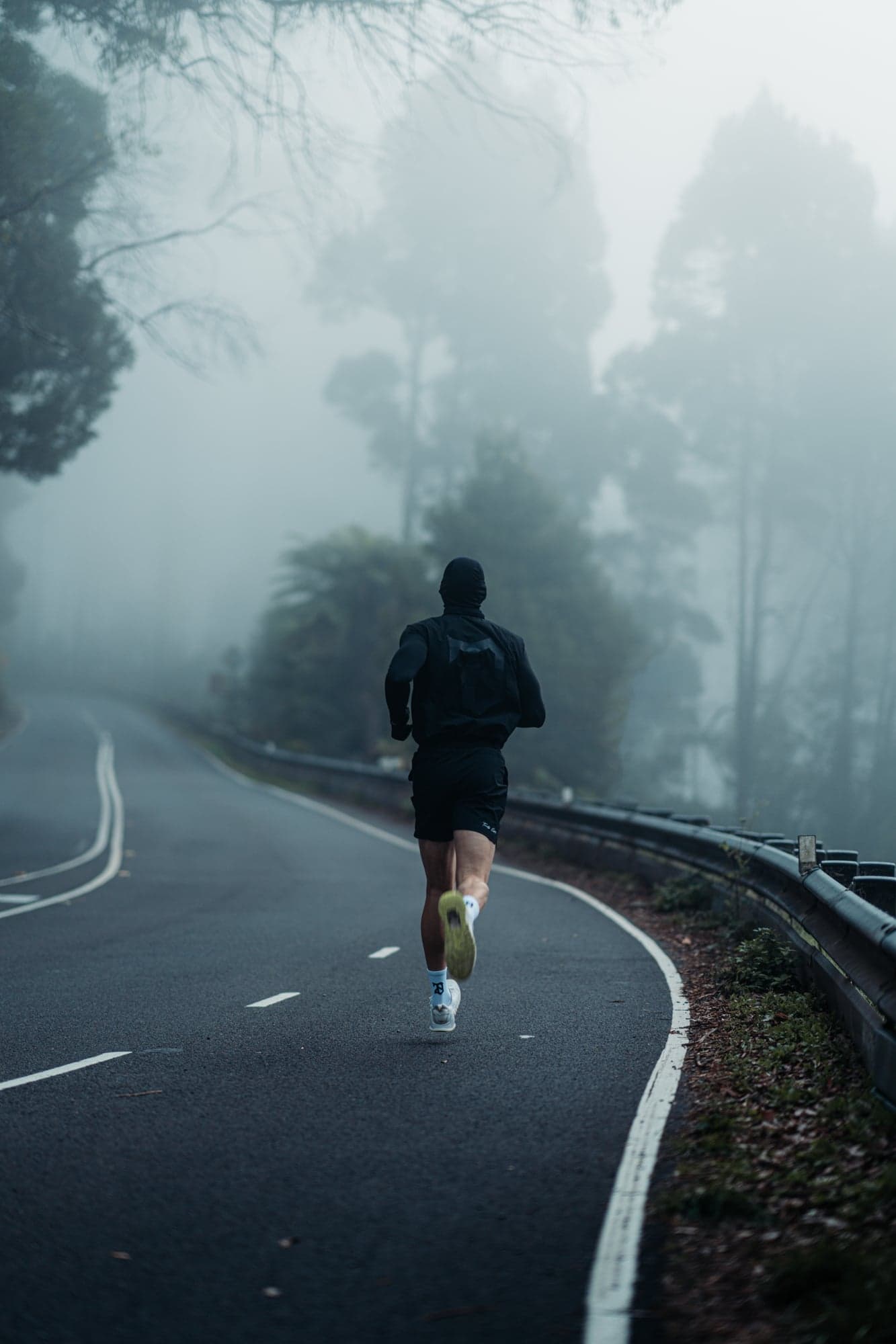 A person dressed in dark athletic wear runs alone on a winding road through a foggy forest, surrounded by tall trees and mist.
