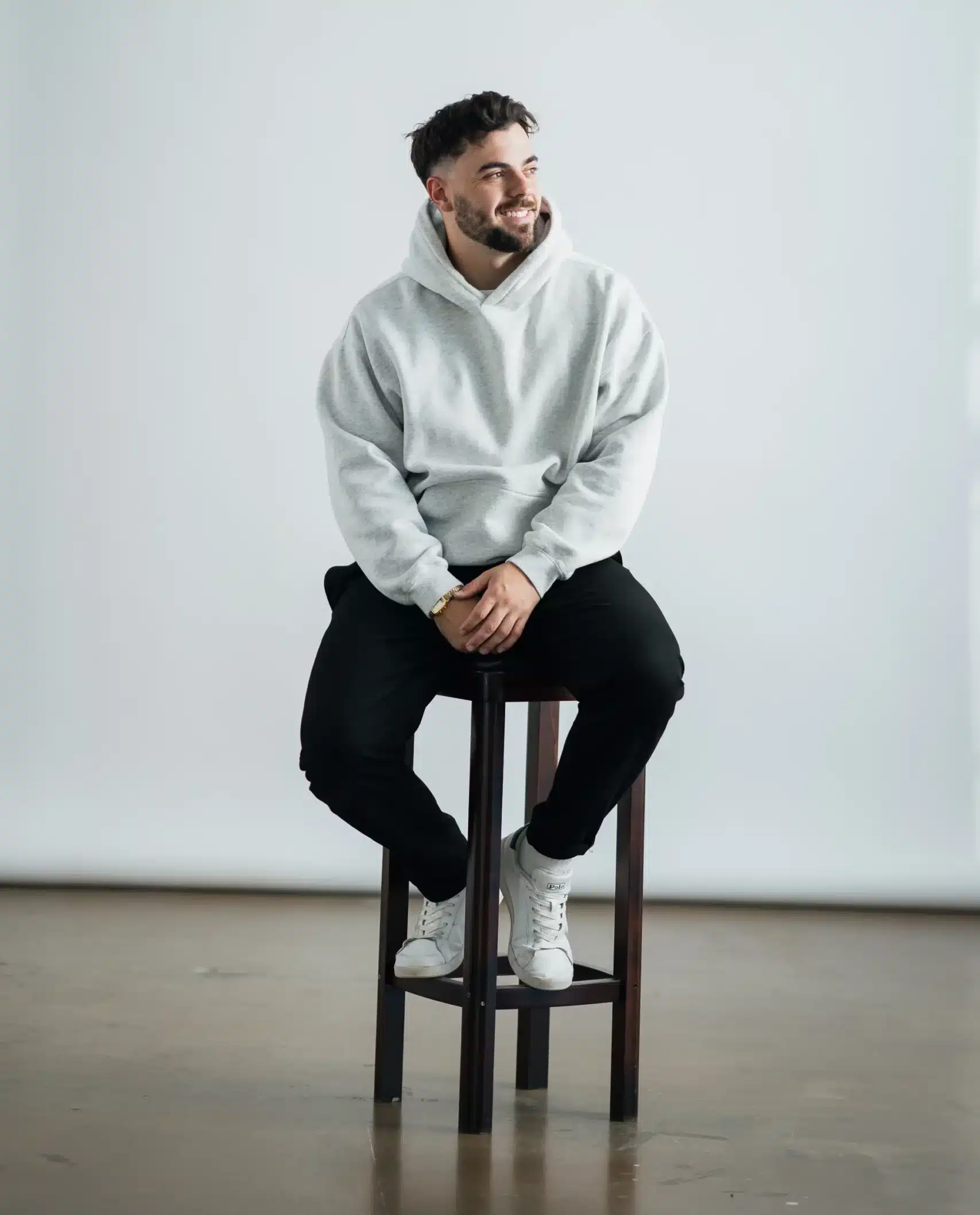 Man with a beard sitting on a stool, wearing a light-colored hoodie and black pants, looking relaxed in a studio setting.