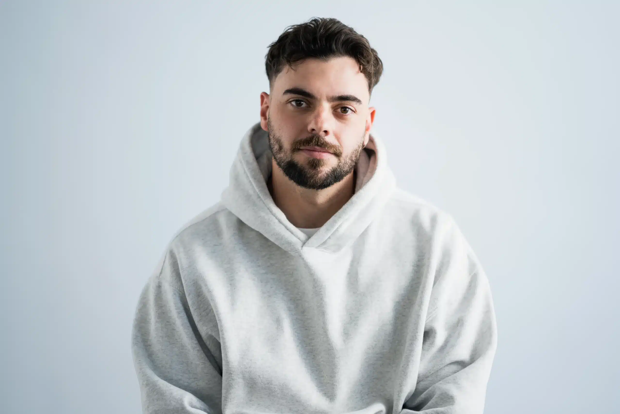 A man with dark hair and a beard wearing a light gray hoodie sits against a plain, light-colored background, looking directly at the camera with a neutral expression.