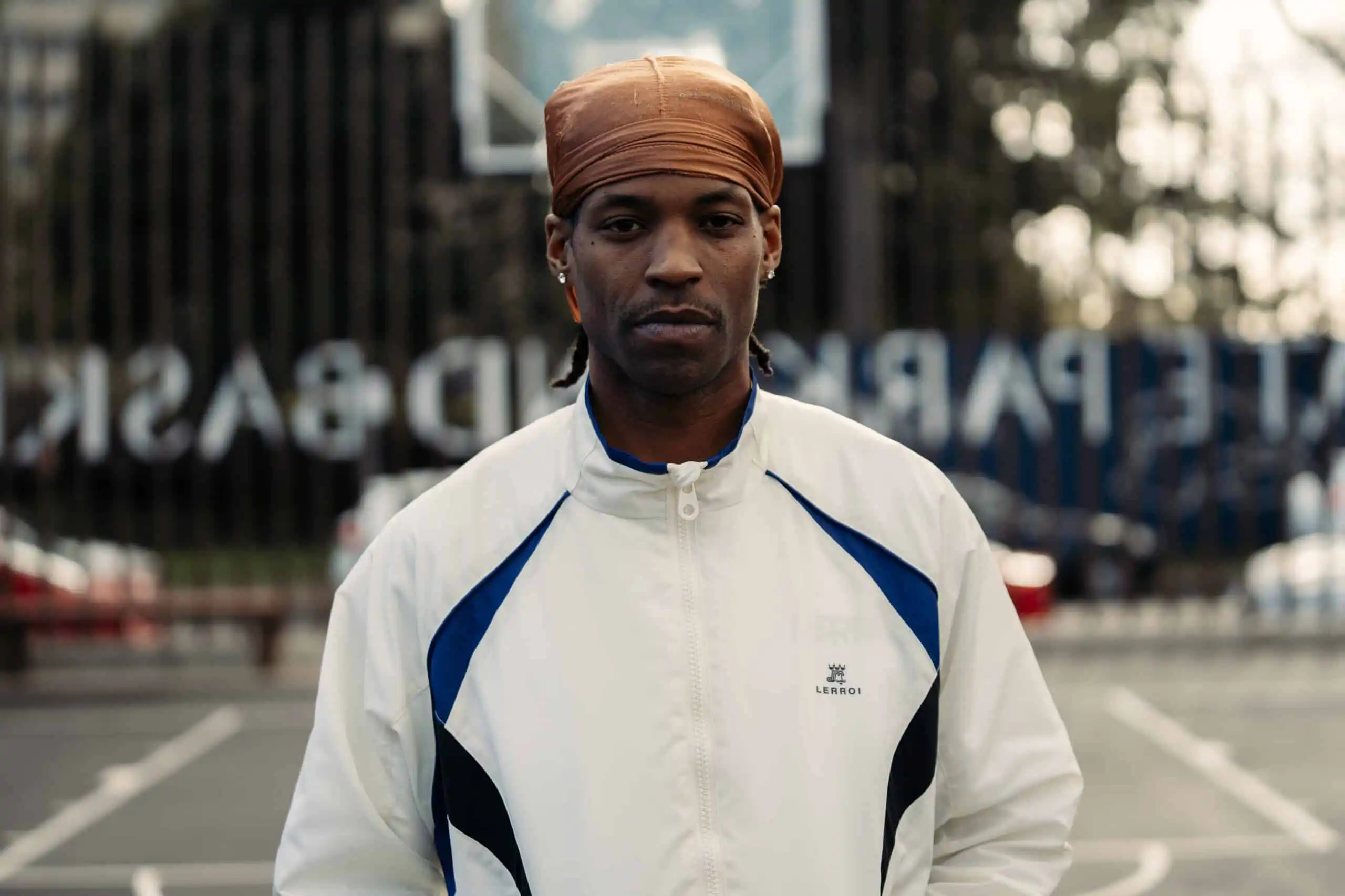 A man wearing a brown headscarf and a white track jacket with blue accents stands on an outdoor basketball court, looking seriously at the camera. The background is blurred, showing a hoop and some trees.