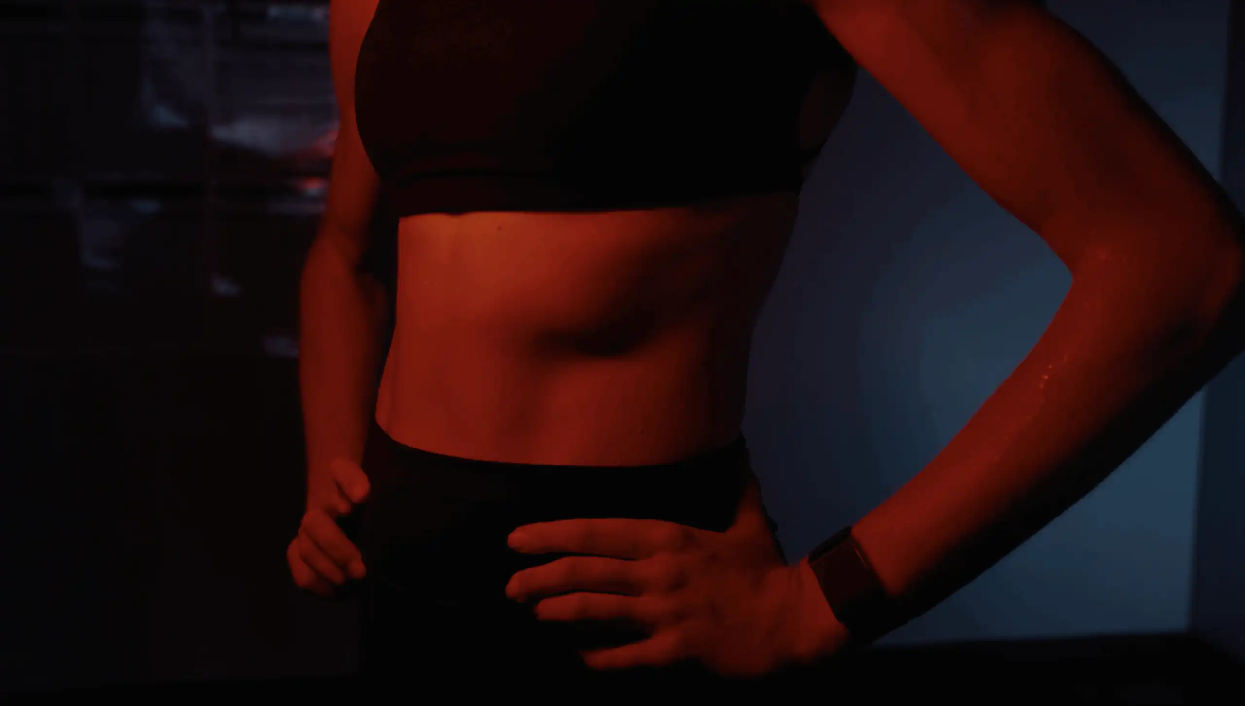 A person wearing a sports bra and fitness watch stands with hands on hips, showing a toned midsection. The lighting is dark with a red and blue tint, creating a dramatic and intense atmosphere.